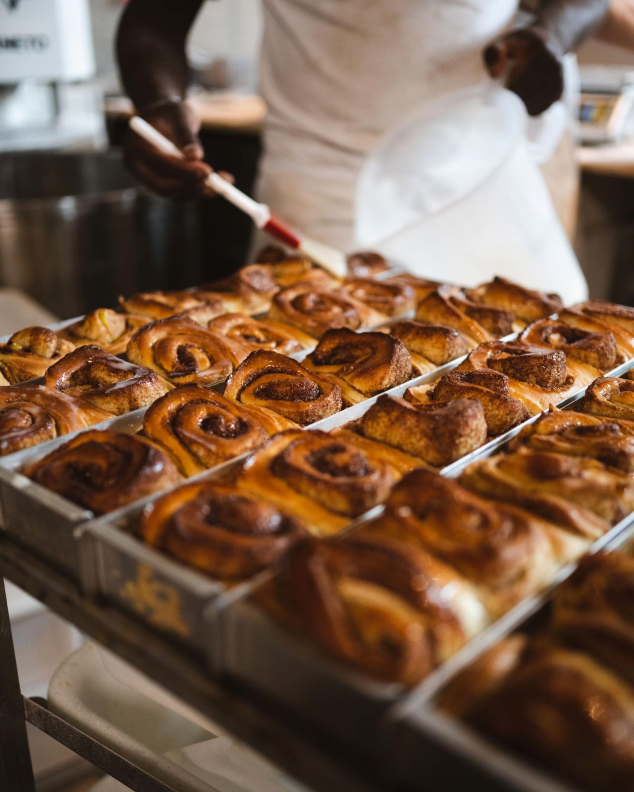 Ein B&auml;cker bestreicht frisch gebackene Zimtschnecken in einer Metallform. Die Zimtschnecken sind goldbraun und gleichm&auml;&szlig;ig angeordnet.