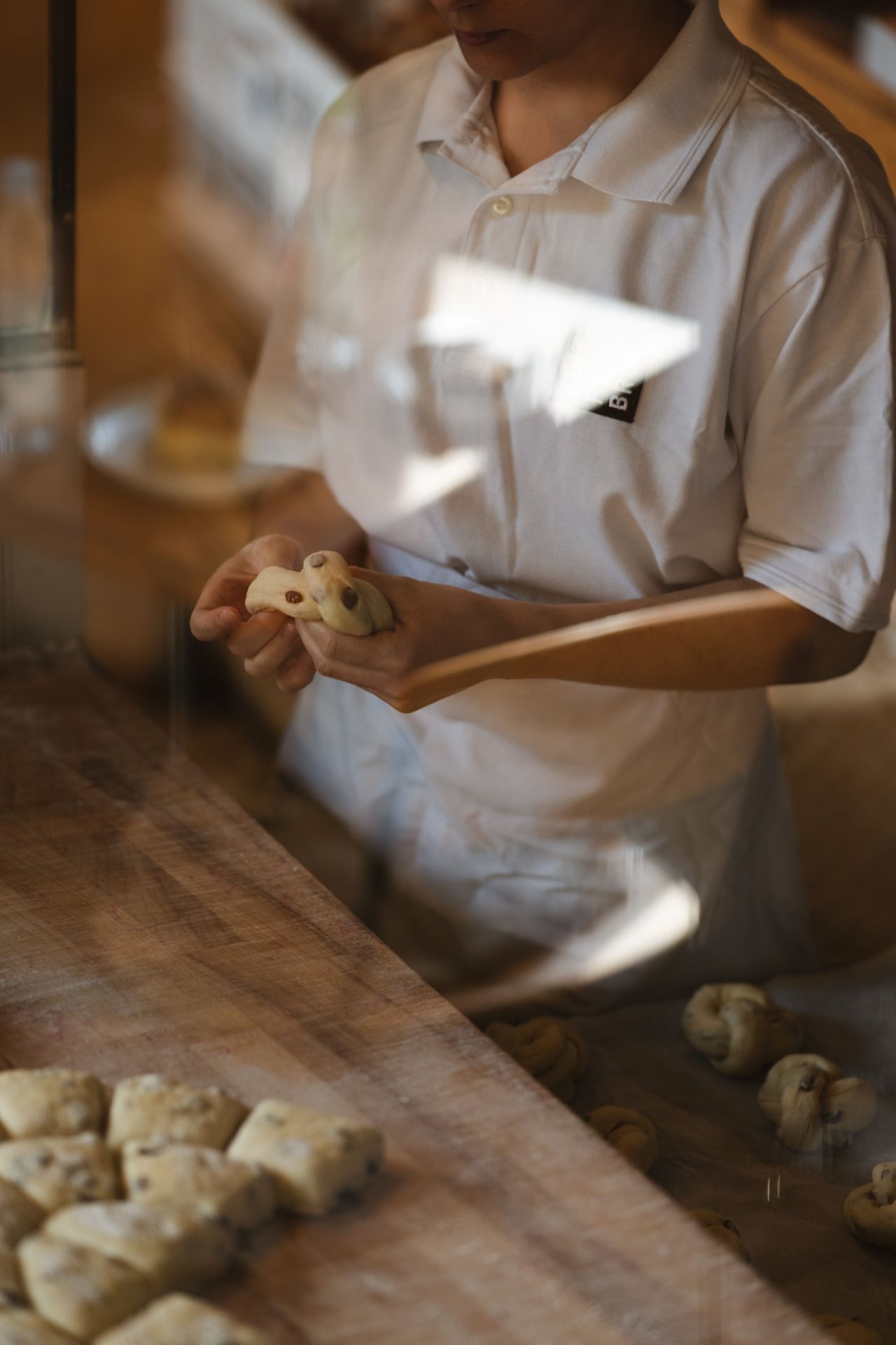 Eine Person in einem wei&szlig;en T-Shirt formt Teig mit beiden H&auml;nden. Der Hintergrund zeigt eine Holzoberfl&auml;che und weitere Teigst&uuml;cke.