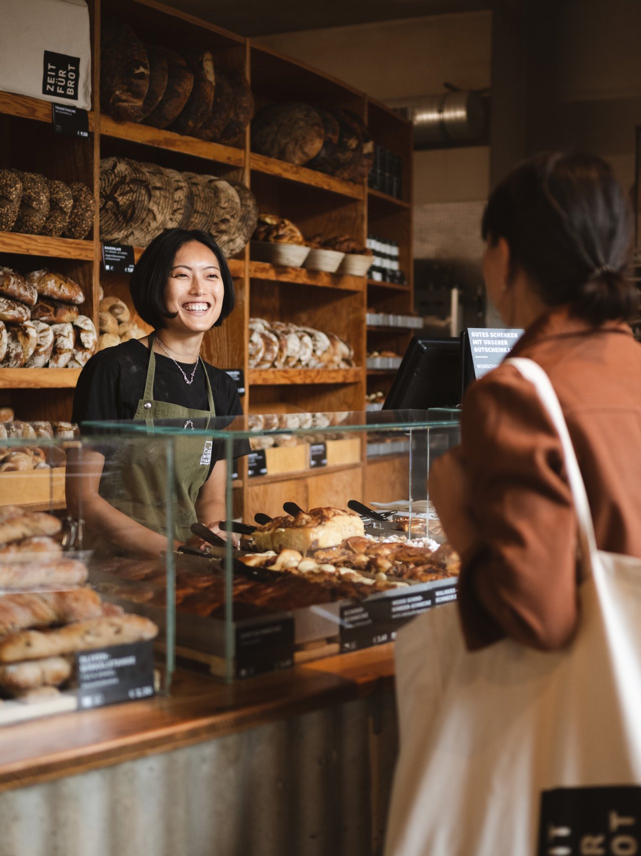 Eine lachende Person steht hinter einer Glastheke, umgeben von verschiedenen Broten und Geb&auml;ck. Sie tr&auml;gt eine Gr&uuml;ne Verkaufssch&uuml;rze. Eine Kundin mit einer hellen Tasche und roter Jacke schaut zur Verk&auml;uferin.